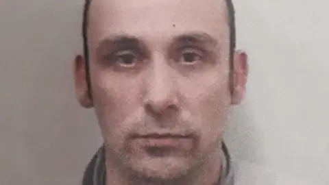 Daniel Washbourne is looking at the camera with a stubbly moustache. He has short brown hair. The image is low-quality.