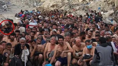 supplied A group of dozens of detained Gazan men pictured in their underwear, during what appears to be an Israeli check for weapons and signs of any links to Hamas. They are sitting or squatting, some young and some older. At the far left of the picture, in the middle of the group, a little girl with dark hair can be seen. The BBC has chosen to blur the faces of the men nearer the front of the picture. In the background is a destroyed building, collapsed into rubble.   