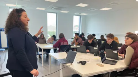 Emma Baugh/BBC Woman in a blue suit at the front of a classroom speaking to students at desks in front of her. The students all have laptops.