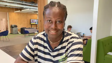 A woman sitting down, wearing a blue and white hooped top. She is also wearing a gold necklace, and on a table behind her are two students sitting down on laptops.