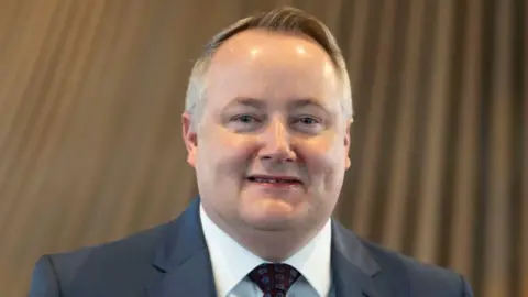 Darren Millar stood in front of the wooden funnel in the Senedd. He is smiling and wearing a grey suit, tie and white shirt.