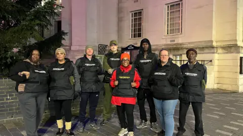 Holly Nichols/BBC A group of eight volunteers are standing together outdoors in front of a large stone building with tall windows and steps leading up to an entrance. The volunteers are wearing black jackets and black stab-proof vests with white text that reads “Street Fathers” or “Street Mothers.” One person in the centre stands out in a bright red coat under the vest.