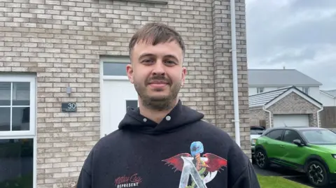 Connor Davidson standing in front of his brick house. he wears a Motley Crue Hoody and has brown hair and stubble. 