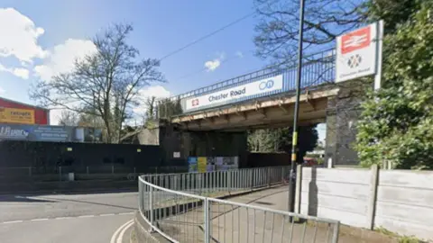 A road with metal railings in the foreground and a bridge overhead with the words Chester Road on it