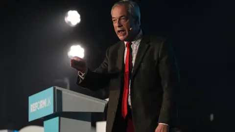 Nigel Farage wearing a brown suit and a red tie stands at a Reform UK podium.