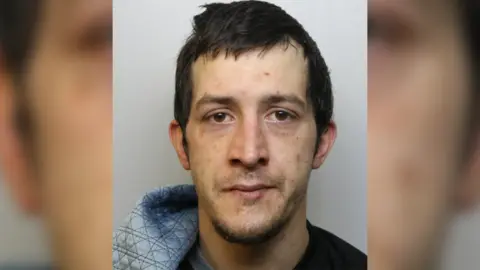 A police photograph of a man with dark hair and brown eyes. He is wearing a hooded top and looking straight at the camera. 