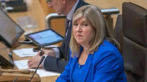 PA Media Woman in a blue suit sits at a desk with a microphone
