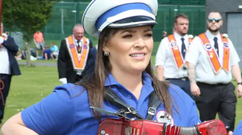Pacemaker Archive photo: A young woman from Portaferry Accordion Band parading in Newtownards on 12 July last year. She is wearing a blue polo shirt with a crest of the band, a white hat with a blue band and a black peak and three silver earrings in her right ear. She is smiling and playing a red accordion. Orangemen are marching on a grassed area behind her.