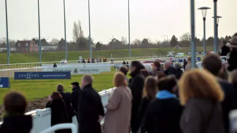 A crowd of people watches on as horses race along the track at Chelmsford City Racecourse.