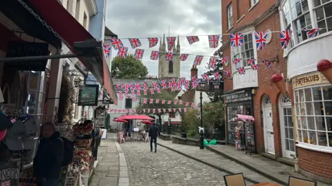 A cobblestone street with historic buildings on either side that now house shops. There is a church in the background and bunting with the union flag has been hung up