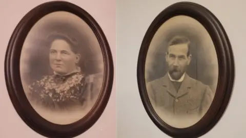 (L-R) May and Alfred Stephan pictured in black and white individual pictures in brown circular frames, hanging on a wall.