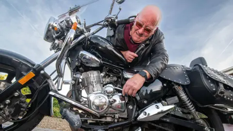 John Peacock leans over his Keeway Superlight bike. He is wearing a leather jacket, sunglasses and a watch on his left wrist.