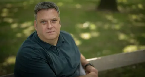 Steven is wearing a blue shirt and is sat on a bench outside. He is in focus but the background, which resembles grass and trees, is fully out of focus. The camera angle is raised and Steven is looking up into the camera with a straight face. He has blue eyes and short grey hair.