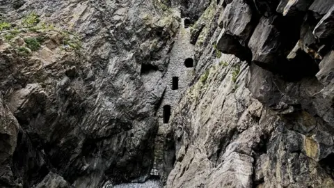 Wide view looking up through a rocky gorge with jagged grey cliff walls on both sides. A tall, narrow stone structure is embedded between the cliffs, with small window openings, and a dry, pebble-covered riverbed lies at the bottom.