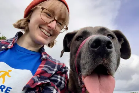 Yorkshire Cancer Research Alice Hildred with short blonde hair wearing an orange hat. She is smiling and is wearing glasses. She has a blue t-shirt, a chequered shirt and is with a brown dog which is sticking its tongue out.