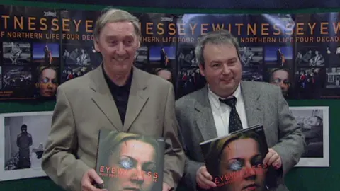 Brendan Murphy wearing a tan blazer and black shirt holding a book with a woman with shattered glass over one eye which is titled Eyewitness. Beside him in Seamus Kelters wearing a grey blazer, white shirt and black tie holding the same book.