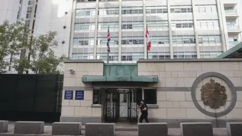 A defender  successful  a achromatic  outfit walks past   the gated entranceway  to the British Embassy, a several-storey precocious   bureau   block, successful  Moscow