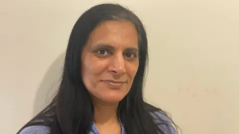 Rashida Anwar Rashida Anwar with long black hair wearing a blue top with a neutral expression standing against a plain cream wall