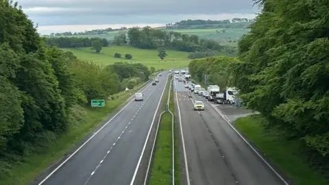 Guy Renner-Thompson Cars are being diverted off a dual carriageway onto a slip road by a police car. Each side of the road has green trees and fields 