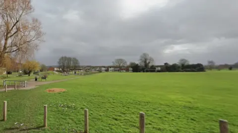 A large green playing fields with houses along two sides. A goal post is visible in the distance and a row of fence posts sit in the foreground. 