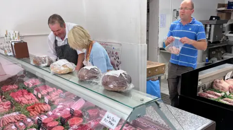 BBC/James Ingham Edward and several other staff behind the meat counter at a farm shop. Various meats like sausages and burgers are behind a glass counter