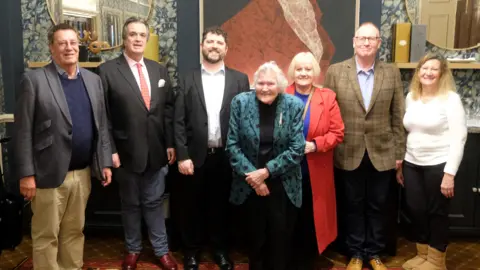 Paul Buchanan A group of seven people in a room, including Gill Green. There are four men, including the Duke of Bedford. The men are all in smart clothes, including jackets. A woman in a red coat is standing behind Mrs Green. Another woman to the right is wearing a white top, black trousers and beige boots. They are all smiling. 