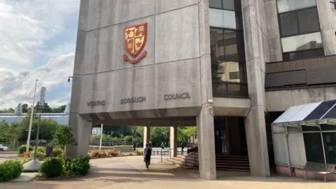 Outside of the civic offices in Woking. A grey concrete building with the words "Woking Borough Council" and a coat of arms above it. Plants and trees can partially be seen in the background. A man walks pass some steps into the building.