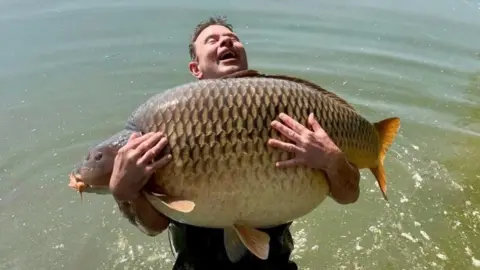 A man looks at the sky with a delighted expression on his face, with his eyes closed, while holding a gigantic fish - a carp - which is grey, gold and yellow in colour