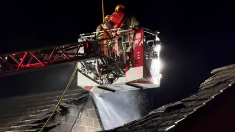 Mid and West Wales Fire Service Two fire fighters seen pouring water into a hole in the roof of a house from a crane