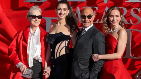 Meryl Streep, Anne Hathaway, Stanley Tucci and Emily Blunt pose on the red carpet in London. Streep is wearing sunglasses, a long, red, satin jacket, a cream blouse and black trousers. Hathaway has her hair up and is wearing a black embellished corset. Tucci is wearing a pinstriped black suit, white shirt and tie, along with sunglasses. Blunt has a red two-piece on an a jewelled necklace.