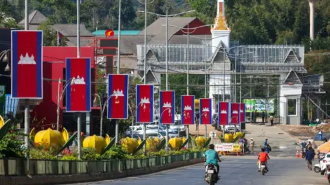 The Ban Pakkad Border Checkpoint between Thailand and Cambodia. People are seen riding two-wheeled vehicles as large banners in the colour of Cambodian flag line the street.t
