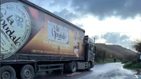 The image shows a large articulated lorry stopped on the side of a rural road. The trailer is wrapped in advertising for a French beer brand, with a big image of a beer bottle cap and the words “La Goudale” and “Les Brasseurs du …” clearly visible along the side. The road surface looks wet, suggesting recent rain, and the sky above is grey with heavy clouds breaking to lighter patches near the horizon.