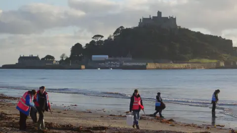 Five people in high-viz jackets litter picking on a beach in front of St Michael's Mount