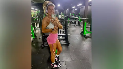 Jane Woodhead Jane in a white top and pinks shorts taking a selfie in a gym