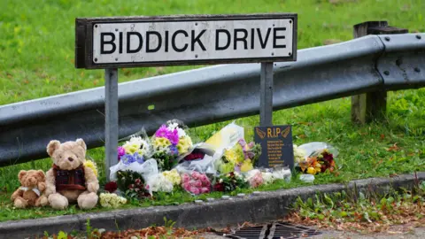 PA Media A sign for Biddick Drive with flowers left underneath. There are two teddy bears on the left on the ground and a small sign saying RIP.