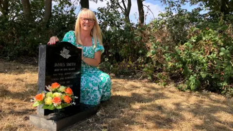 Una mujer de mediana edad, con cabello rubio largo y liso, gafas y un vestido turquesa con estampado de hojas blancas, se agacha junto a una lápida de mármol oscuro grabada con el nombre de James Smith. Justo detrás de ella hay una hilera de árboles y arbustos.