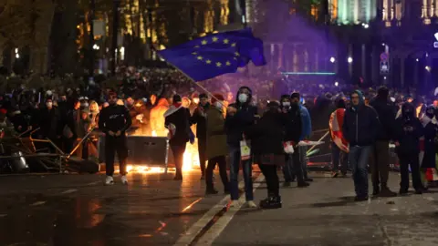 Getty Images Georgian police use water cannons and tear gas to disperse protesters in Tbilisi. A man can be seen waving a European Union blue and yellow star flag against the backdrop of a fire blazing and large crowds of protests