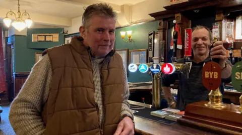 A man wearing a brown gilet and mottled grey jumper leaning on a brown wooden bar with a second man behind the bar holding on to a brass-topped, wooden handled beer pump.