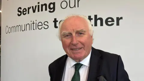 Martin Heath/BBC Llew Monger wearing a dark suit, white shirt and green tie stands indoors in front of a white wall displaying the words “Serving our Communities together”. The image is a head-and-shoulders portrait taken in a modern office or public building setting.