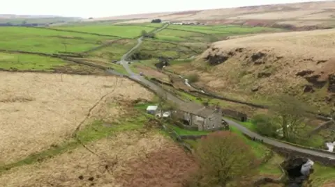 An area of land - mostly fields, with a road running through the middle and a farm house on one side of the road