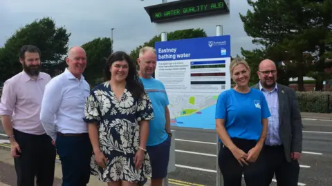 Portsmouth City Council Various councillors and local activists stand in front of a blue sign titled 'Eastney bathing water' with a road behind them. Above the sign, there's an electronic display saying 'no water quality alert' in green letters.
