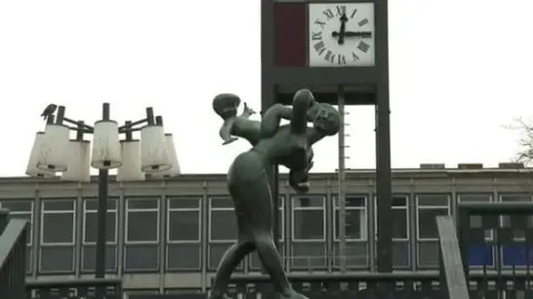 The centre of Stevenage, showing the top floor of a concrete office block with a sculpture in front of it featuring a woman carrying a child. There is a clock above the sculpture, and a lighting column to the left with bulbs enclosed by glass shades.