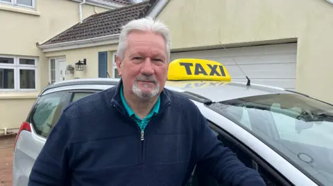 Mick Tostevin wears a blue jumper with a turquoise shirt underneath. He's leaning on his silver taxi, outside a house.