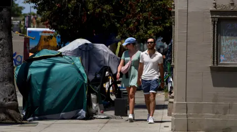 EPA A man and woman walk on a sidewalk in Los Angeles as the woman peers over at several tents where homeless people are residing. 