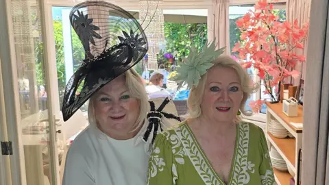 Paula Gamester Paula Gamester and a friend standing in her conservatory in the sunshine. both are blonde and Paula is wearing a cream dress with a large wired black hat with flowers in the pattern. Her friend is wearing a sage green dress with white embroidery and a floral fascinator