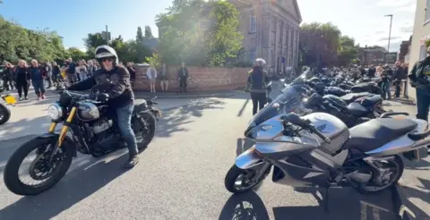 BBC To the right of the photo are a row of motorbikes all lined up on the road. People are gathered around to look at them. To their right is a man who is riding a black motorbike. He is wearing a black jacket and black helmet. 