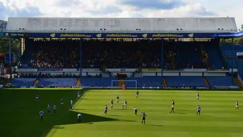 Getty Images A straight on view of a stand in a football stadium. It is one of the smaller stands (along the shorter side of the pitch) and the seating is blue. It is a sunny day and some of the players on the pitch cast shadows.