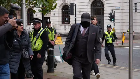 BBC Robert Bush arrives at Hull Crown Court in a black suit wearing a cap and face mask he is surrounded by police officers and press