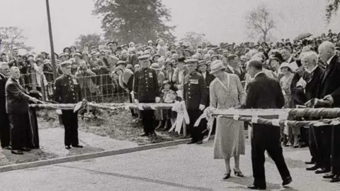 A black and white photo of a woman in light dress and hat, cutting a ribbon on road barriers