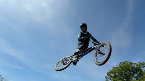 Connor Bennett/BBC A cyslist is caught in a mid-air jump against a blue sky